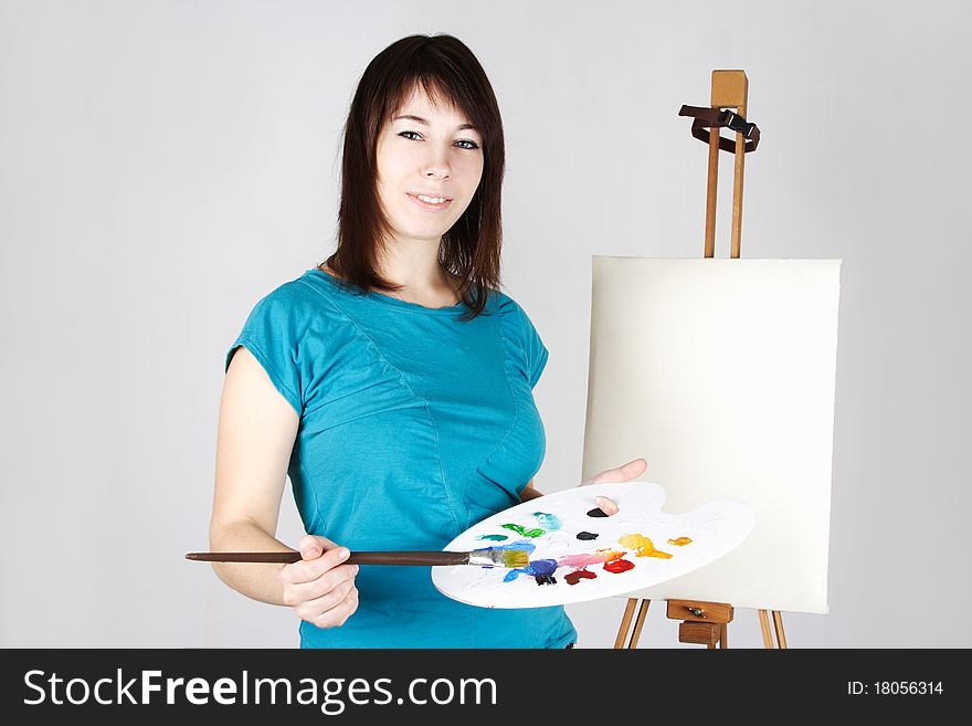 Young girl in blue shirt standing near easel, holding brush and palette, smiling. Young girl in blue shirt standing near easel, holding brush and palette, smiling