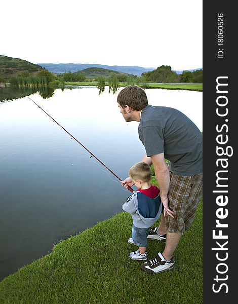 A father is teaching his young son to fish in a peaceful lake surrounded by green trees and grass. A father is teaching his young son to fish in a peaceful lake surrounded by green trees and grass.