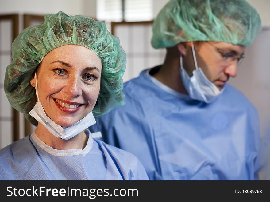 A lovely nurse with her surgical mask away from her face smiles at the camera with an out of focus doctor in the background. A lovely nurse with her surgical mask away from her face smiles at the camera with an out of focus doctor in the background.