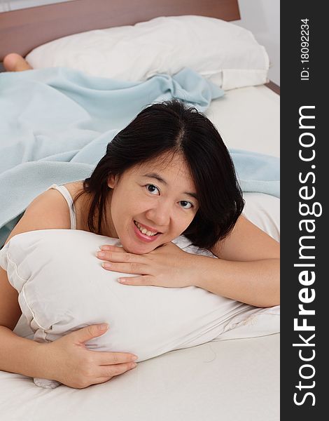 A smiling Asian woman in bed hugging a pillow while looking at you