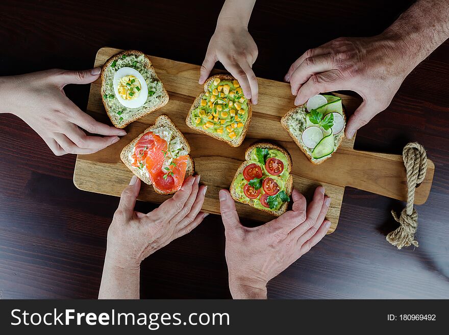 Many hands of different ages of people reach out and hold various healthy and delicious sandwiches. Slices of bread are spread with cottage cheese and avocado sauce, various vegetables on top. Many hands of different ages of people reach out and hold various healthy and delicious sandwiches. Slices of bread are spread with cottage cheese and avocado sauce, various vegetables on top