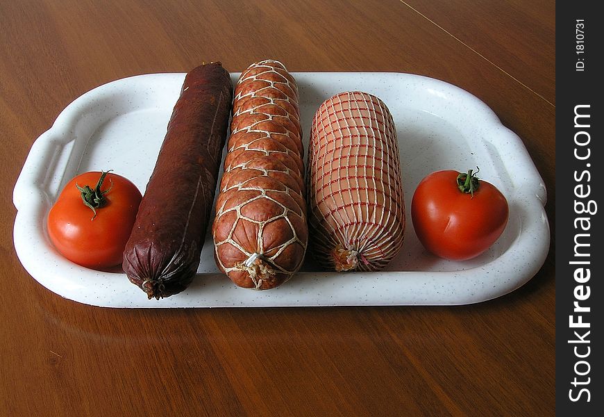 On a photo on a white tray 3 kinds of sausages, and from both sides two red tomatoes. The photo is made in Ukraine. On a photo on a white tray 3 kinds of sausages, and from both sides two red tomatoes. The photo is made in Ukraine.