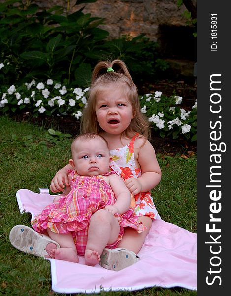 Two very young girls sitting on a blanket in a park making very grumpy faces. Two very young girls sitting on a blanket in a park making very grumpy faces.