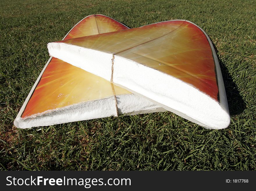 A Broken Yellow, Orange Surfboard On A Green Grass Field, Result Of Dangerous Surfing Conditions. A Broken Yellow, Orange Surfboard On A Green Grass Field, Result Of Dangerous Surfing Conditions