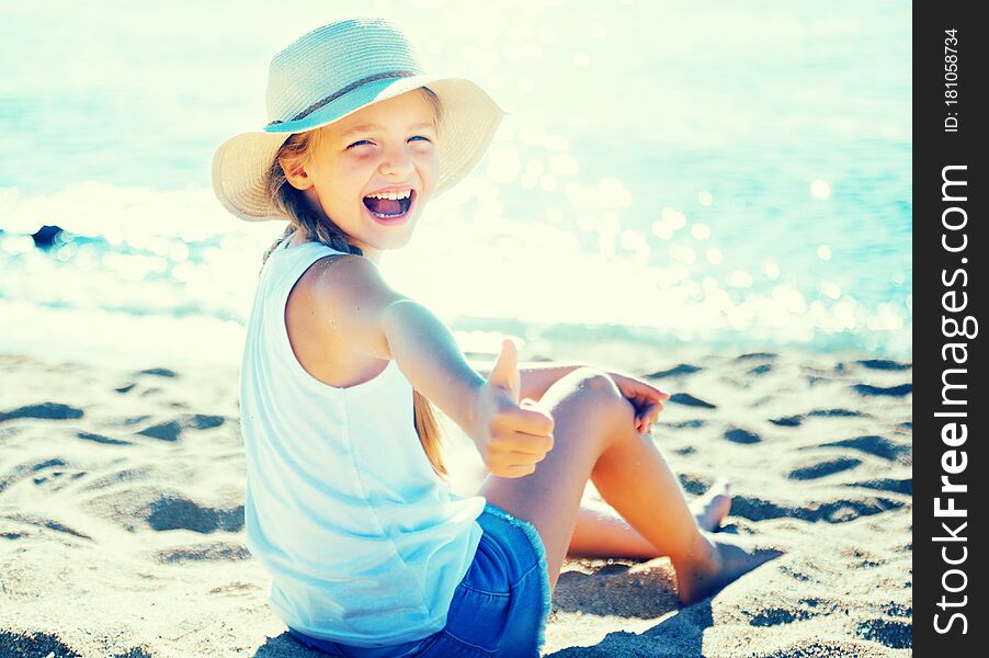 Blond child in hat on beach shows thumb up