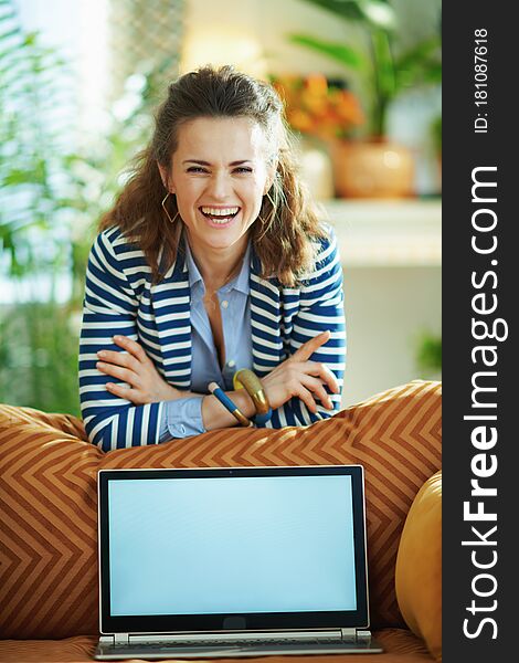 Portrait of smiling trendy 40 years old woman in blue blouse and striped jacket at modern home in sunny day near couch showing laptop blank screen. Portrait of smiling trendy 40 years old woman in blue blouse and striped jacket at modern home in sunny day near couch showing laptop blank screen