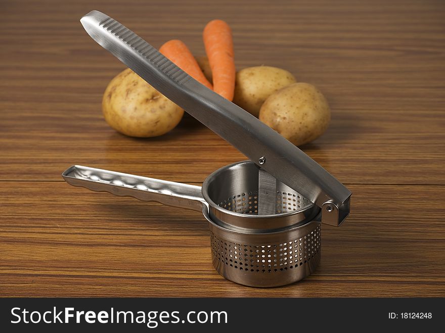 A horizontal image of a food ricer masher and vegetables on a wooden table. A horizontal image of a food ricer masher and vegetables on a wooden table