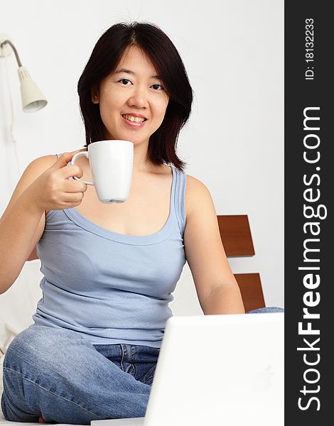 An Asian woman having a cup of coffee while working with a laptop. An Asian woman having a cup of coffee while working with a laptop