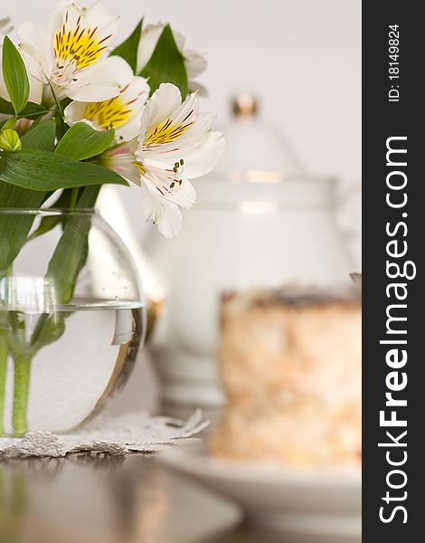 Delicate flower in glass vase on wooden table, teapot and cake on foreground