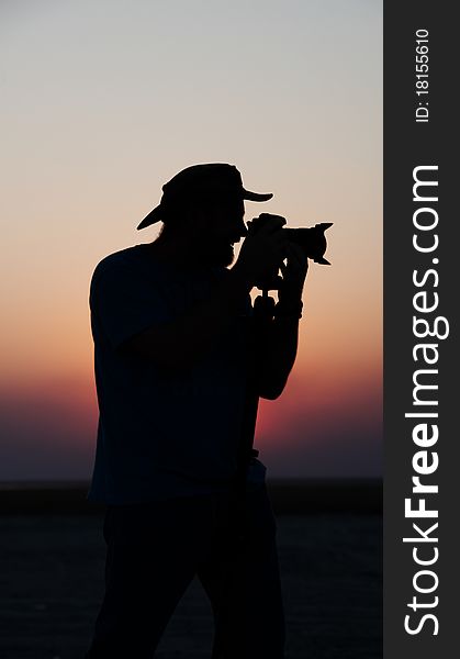 Silhouette of a young man in a hat taking pictures at sunset using a tripod. Silhouette of a young man in a hat taking pictures at sunset using a tripod