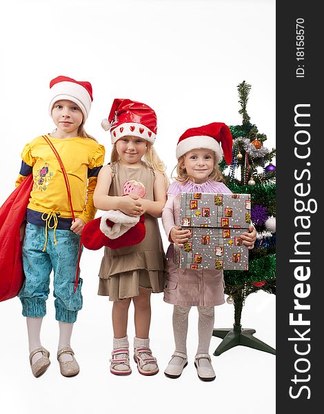 Three young little caucasian blond girls standing together near christmas tree holding gifts and smiling sincerely.isolated over white background. Three young little caucasian blond girls standing together near christmas tree holding gifts and smiling sincerely.isolated over white background