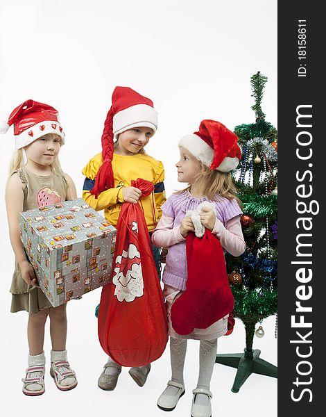 Three young little caucasian blond girls standing together near christmas tree holding gifts and smiling sincerely.isolated over white background. Three young little caucasian blond girls standing together near christmas tree holding gifts and smiling sincerely.isolated over white background