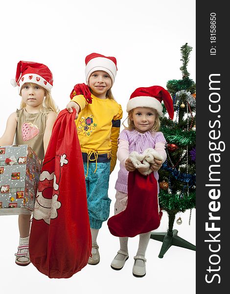 Three young little caucasian blond girls standing together near christmas tree holding gifts and smiling sincerely.isolated over white background. Three young little caucasian blond girls standing together near christmas tree holding gifts and smiling sincerely.isolated over white background