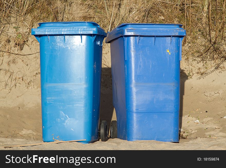 Two blue wheelie bins standing side by side on sand with grasses in the background. Two blue wheelie bins standing side by side on sand with grasses in the background.