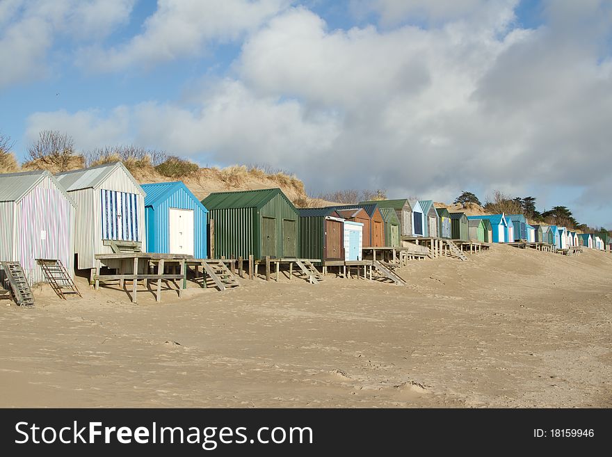 A row of beachhuts, multicoloured, with steps and boardwalks on a beach backed by a sand dune, blue sky and clouds. A row of beachhuts, multicoloured, with steps and boardwalks on a beach backed by a sand dune, blue sky and clouds.
