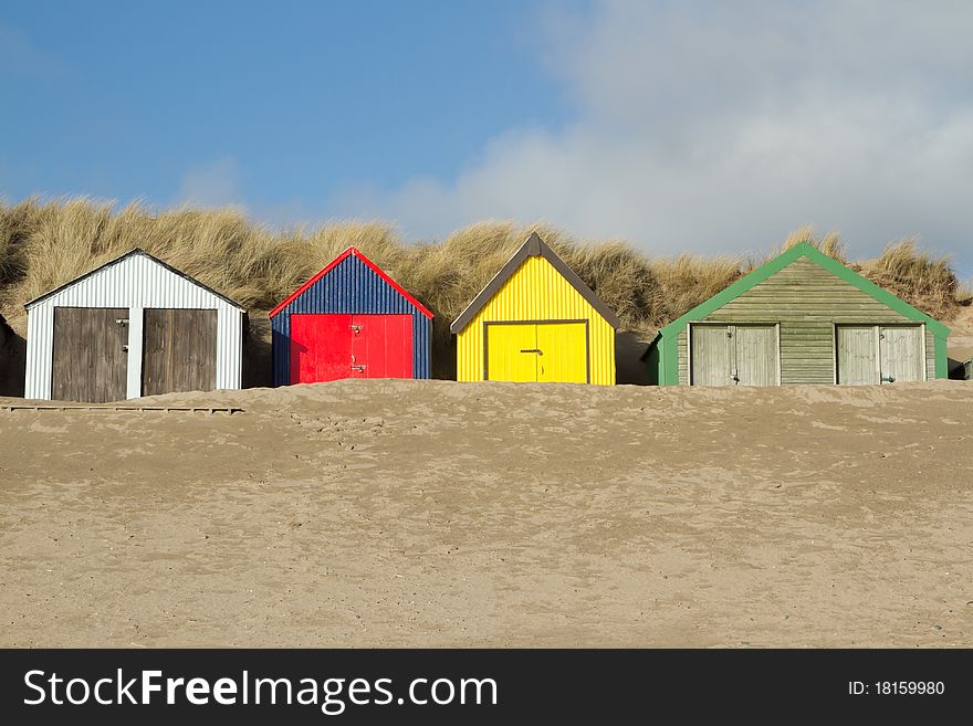 Multi-coloured beach huts with sand banked up against the doors, the scene is backed by a dune with blue sky and cloud. Multi-coloured beach huts with sand banked up against the doors, the scene is backed by a dune with blue sky and cloud.