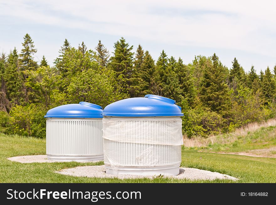 Large garbage and recycling bins sit outdoors in a park. Large garbage and recycling bins sit outdoors in a park.