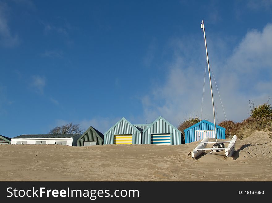 Small yacht, catamaran, in front of beach huts banked up with sand, a blue sky with cloud above. Small yacht, catamaran, in front of beach huts banked up with sand, a blue sky with cloud above.