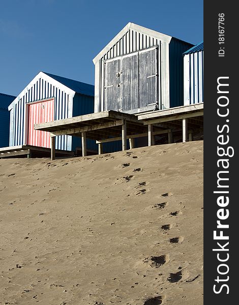Footprints lead up a sand dune to beach huts on raised platforms, a blue sky backs the scene. Footprints lead up a sand dune to beach huts on raised platforms, a blue sky backs the scene.