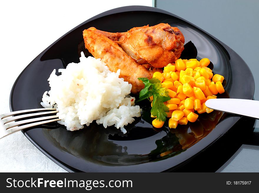 A piece of roast chicken with rice and corn on the black plate. A piece of roast chicken with rice and corn on the black plate