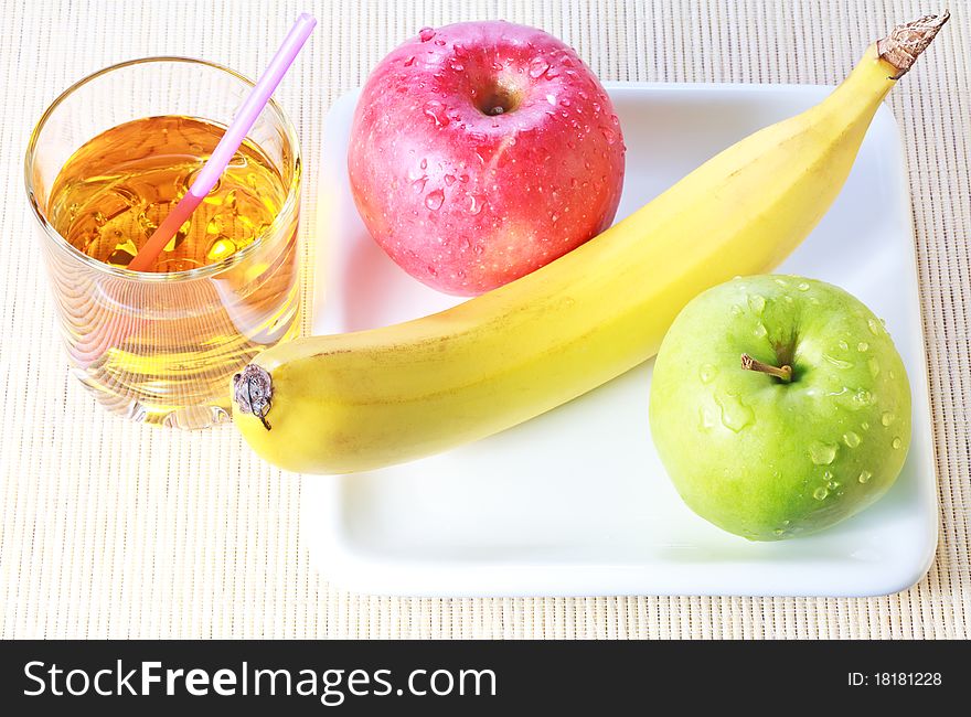 Banana, green and red apples on white plate and glass of apple juice over bamboo mat background. Banana, green and red apples on white plate and glass of apple juice over bamboo mat background