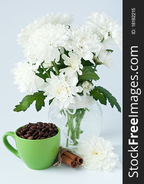 Coffee beans and cinnamon stick in a green cup stands near the glass vase with white flowers