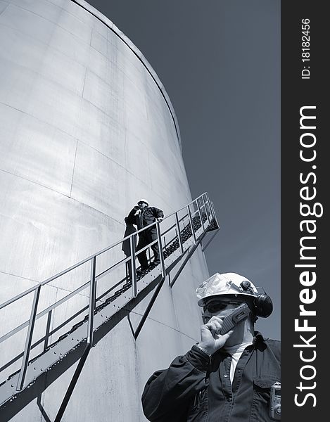 Two oil workers with industrial fuelstorage tanks in background, one standing on stairs. Two oil workers with industrial fuelstorage tanks in background, one standing on stairs