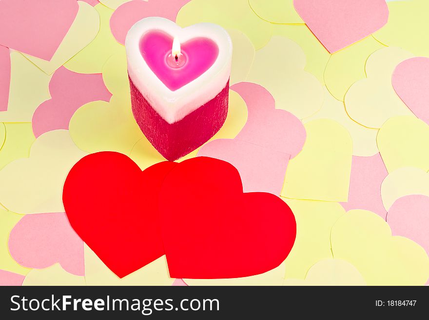 A heart shaped lighted candle over cute paper hearts background and two red hearts in the foreground. A heart shaped lighted candle over cute paper hearts background and two red hearts in the foreground.