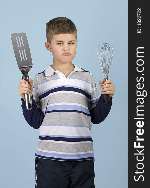 Young boy holding kitchen utensils with an angry expression on a blue background. Young boy holding kitchen utensils with an angry expression on a blue background.