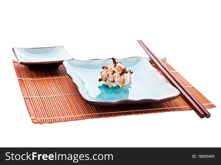 Traditional Japanese sushi tableware set: chopsticks on a rest, plate with a sea shell on it and a saucer, all on a bamboo place mat, on a white background, isolated. Traditional Japanese sushi tableware set: chopsticks on a rest, plate with a sea shell on it and a saucer, all on a bamboo place mat, on a white background, isolated.