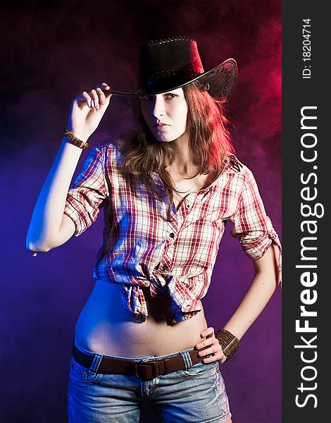 Studio portrait of a young pretty cowgirl in a checkered shirt, jeans shorts and black leather hat on a color smoky background. Studio portrait of a young pretty cowgirl in a checkered shirt, jeans shorts and black leather hat on a color smoky background