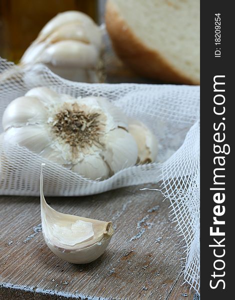 Clove and head of garlic in netting; more garlic, olive oil and bread in background. Clove and head of garlic in netting; more garlic, olive oil and bread in background