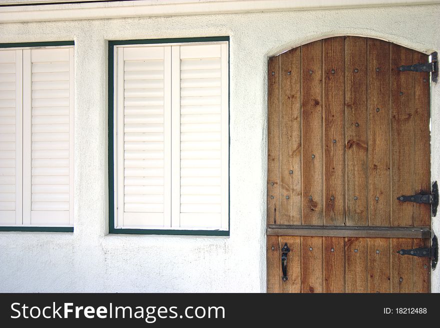 Stained wooden door with black hinges next to white painted shutters and green trim in a stucco wall. Stained wooden door with black hinges next to white painted shutters and green trim in a stucco wall