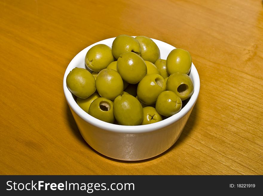 A small white china bowl on a wooden table containing green olives