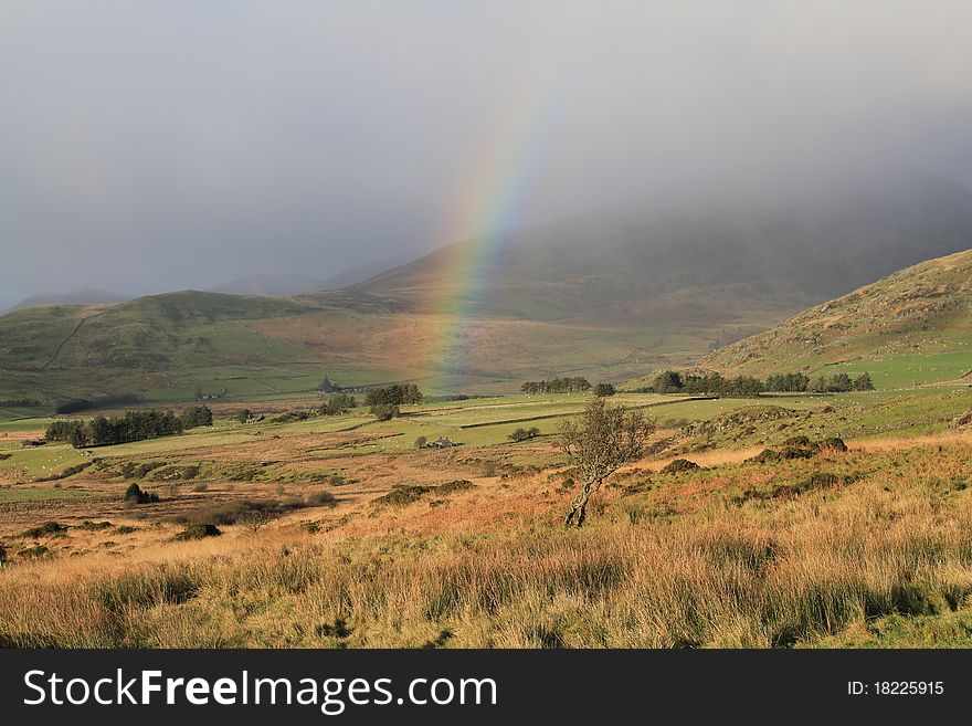 A rainbow forms under a rain cloud in rural fields and trees surrounded by hills. A rainbow forms under a rain cloud in rural fields and trees surrounded by hills.