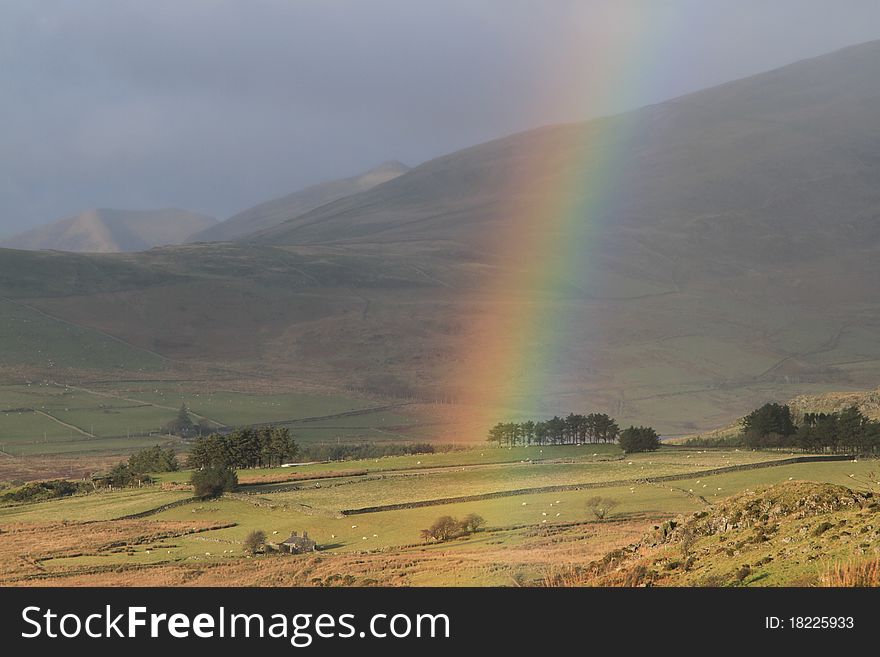 A rainbow forms under a rain cloud in rural fields and trees surrounded by hills. A rainbow forms under a rain cloud in rural fields and trees surrounded by hills.