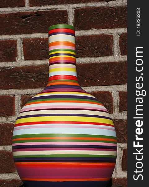 A mulit-colored vase is set against a brick wall. The vase is full of color, including green, red, purple, yellow, white, blue, orange and pink. A mulit-colored vase is set against a brick wall. The vase is full of color, including green, red, purple, yellow, white, blue, orange and pink.