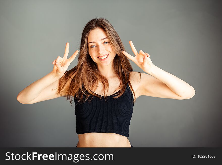 Happy girl in a black top with her hair showing both hands shows a victory sign. Two fingers. Number two. On a gray background. Without makeup, without retouching. Happy girl in a black top with her hair showing both hands shows a victory sign. Two fingers. Number two. On a gray background. Without makeup, without retouching