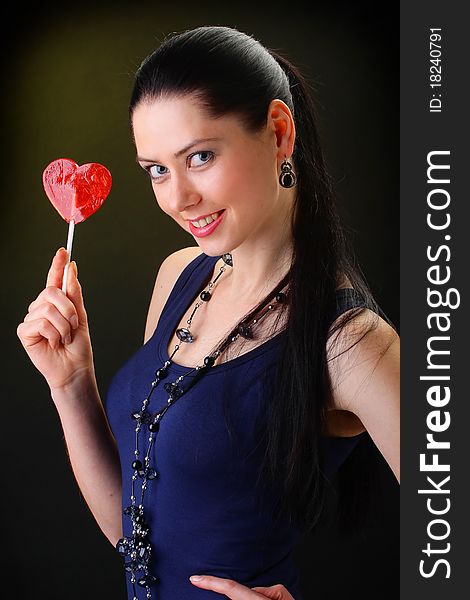 Portrait beautiful young sexy brunette girl with red lollipop heart. On a black background.