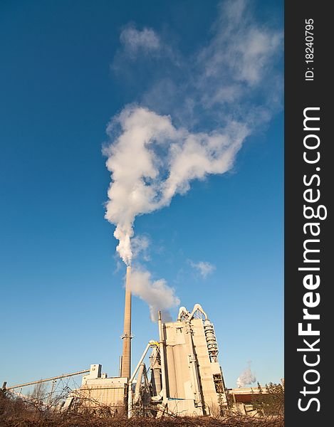 A manufacturing facility pumps smoke into the air from two smokestacks with a clear blue sky in the background. A manufacturing facility pumps smoke into the air from two smokestacks with a clear blue sky in the background.