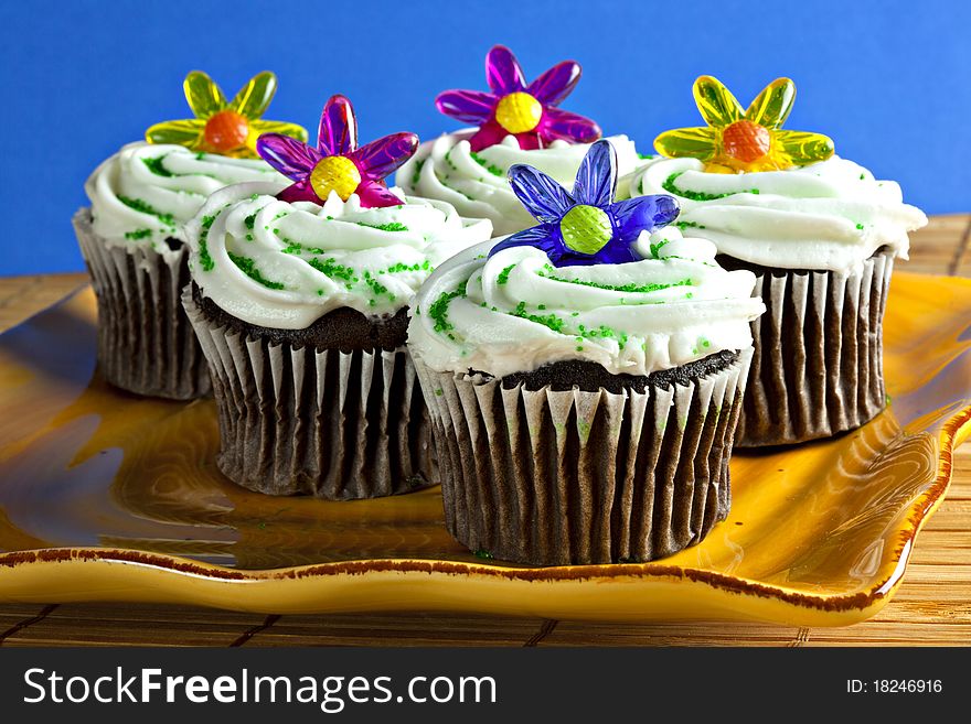 Chocolate Cupcakes with vanilla frosting, sprinkles and a plastic flower on top on yellow plate with blue background. Chocolate Cupcakes with vanilla frosting, sprinkles and a plastic flower on top on yellow plate with blue background