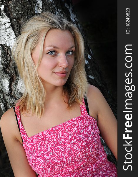 Portrait of a beautiful blonde woman with long hair and a smile near tree in pink dress. Portrait of a beautiful blonde woman with long hair and a smile near tree in pink dress