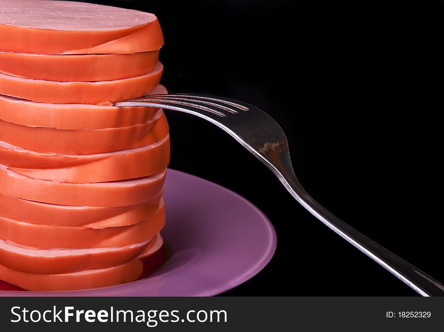 Sliced cooked sausage on the plate with fork across black