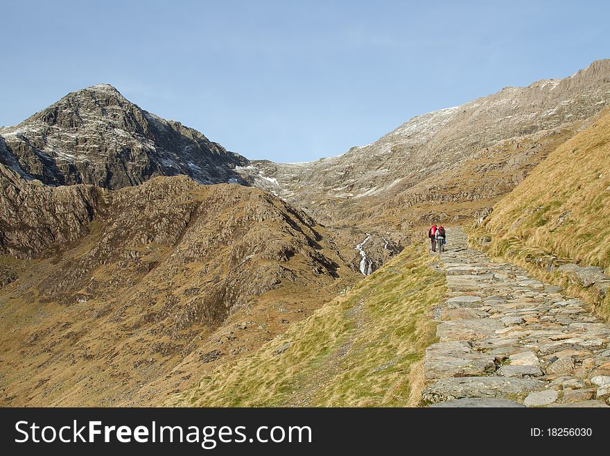 Two people walk up a stone footpath towards a high rocky peak. Two people walk up a stone footpath towards a high rocky peak.