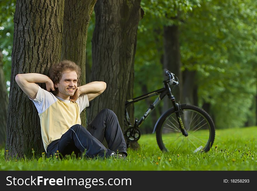 Young man siting in the park under the tree near his bycicle. Young man siting in the park under the tree near his bycicle