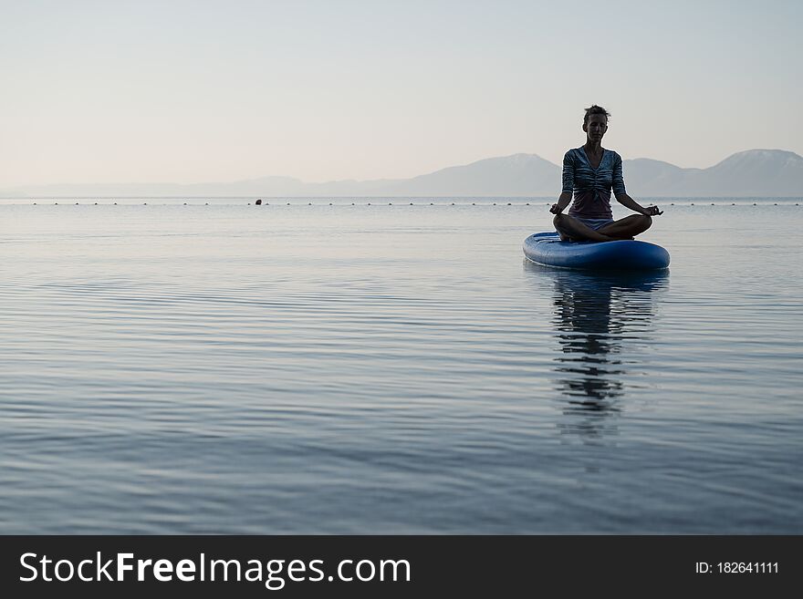 Peaceful young woman sitting in lotus position on sup board floating on calm sea water in early morning. Peaceful young woman sitting in lotus position on sup board floating on calm sea water in early morning