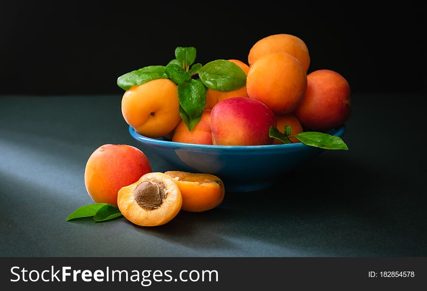 still life, ripe apricots with green foliage in a blue bowl in the rays of sunlight on a dark background