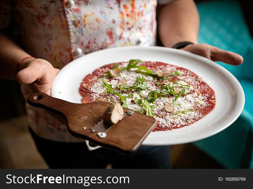 Carpaccio with parmesan, truffles and arugula on a white plate. On a wooden table. 
Chef holds in his hands