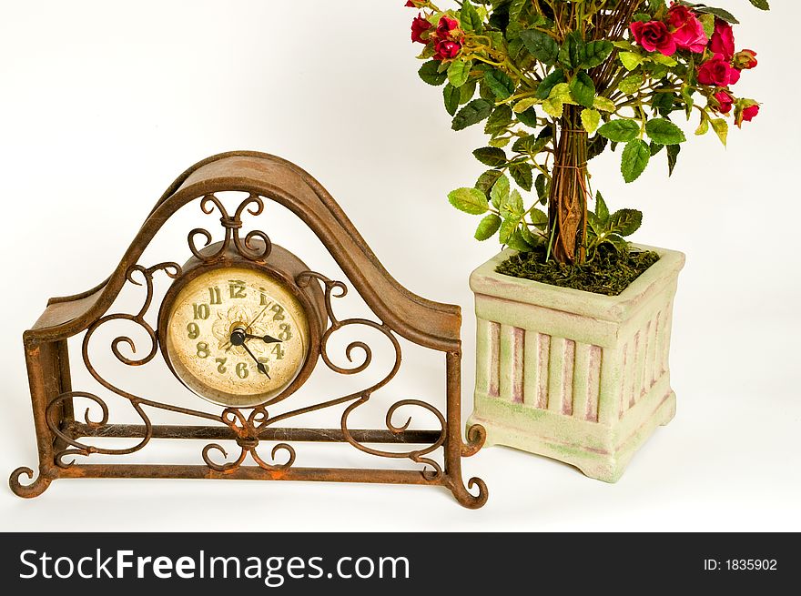 A rusty and dusty old wire-frame clock sits in front of a decorative pot of miniature red roses on a white background. A rusty and dusty old wire-frame clock sits in front of a decorative pot of miniature red roses on a white background.