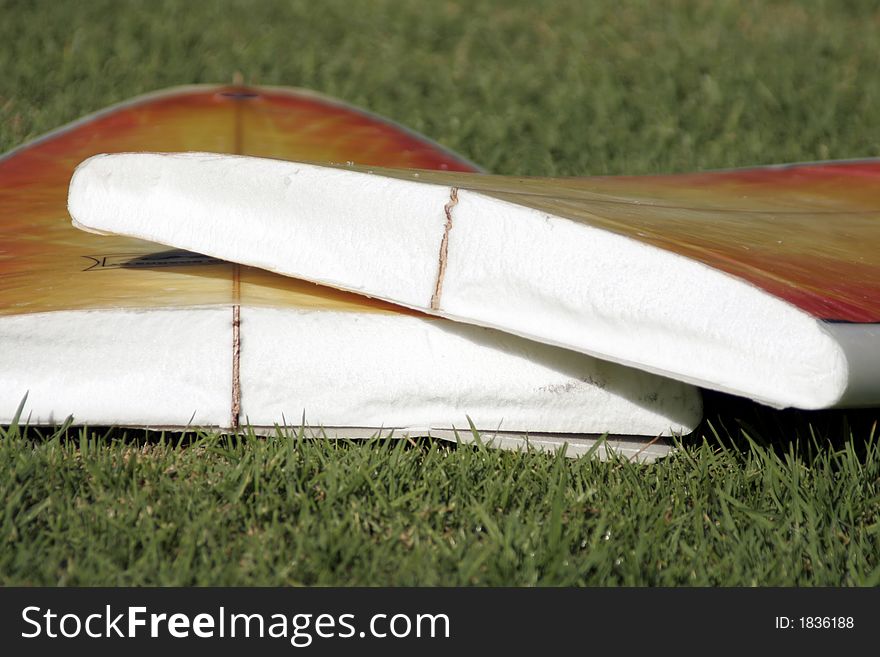 A Broken Yellow, Orange Surfboard On A Green Grass Field, Result Of Dangerous Surfing Conditions, Shallow Depth Of Field. A Broken Yellow, Orange Surfboard On A Green Grass Field, Result Of Dangerous Surfing Conditions, Shallow Depth Of Field